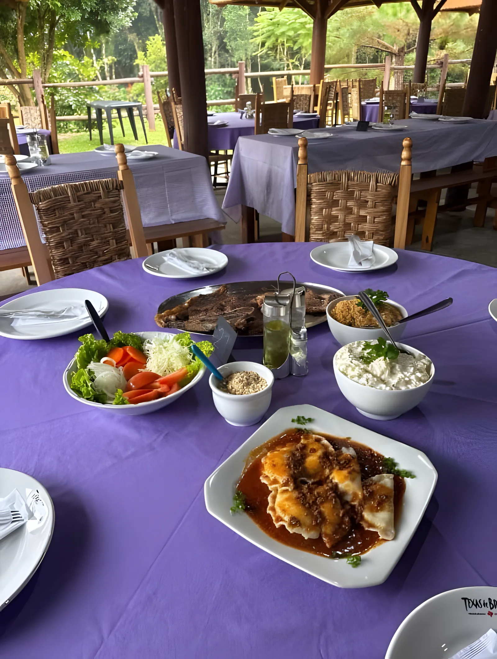 Mesa redonda com toalha lilás servida com churrasco, saladas, maionese e pierogi